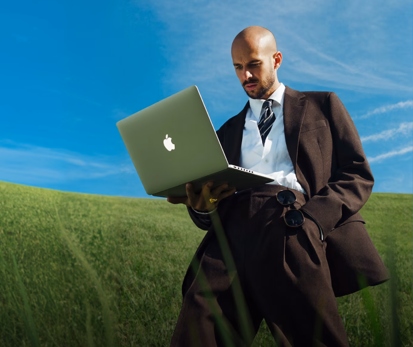A bald man dressed in a dark suit, white shirt, and striped tie stands in a green field holding an open Apple laptop, looking at the screen with concentration under a clear blue sky.