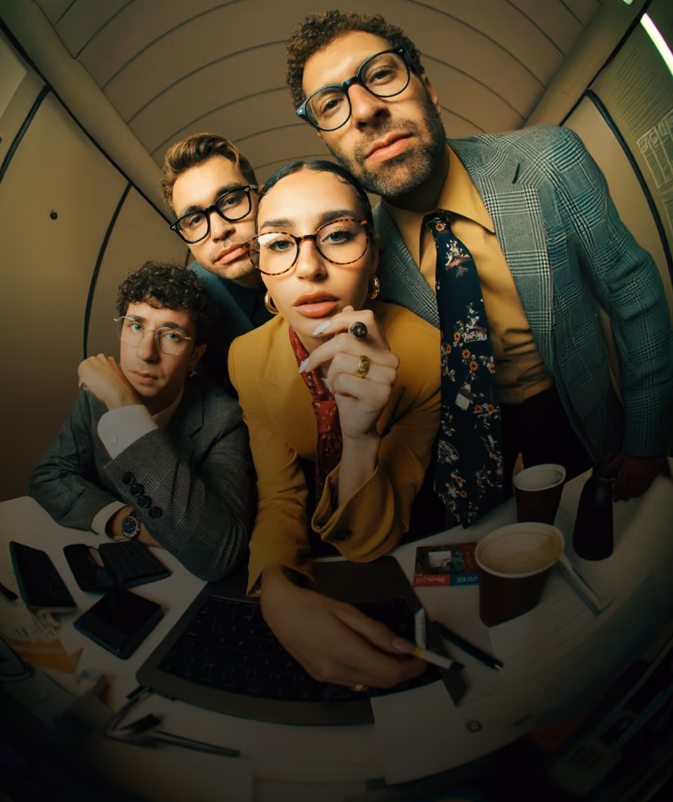 Four young adults wearing glasses leaning closely towards the camera with serious expressions, seated around a table with coffee cups, a laptop, and phones, under a curved ceiling.