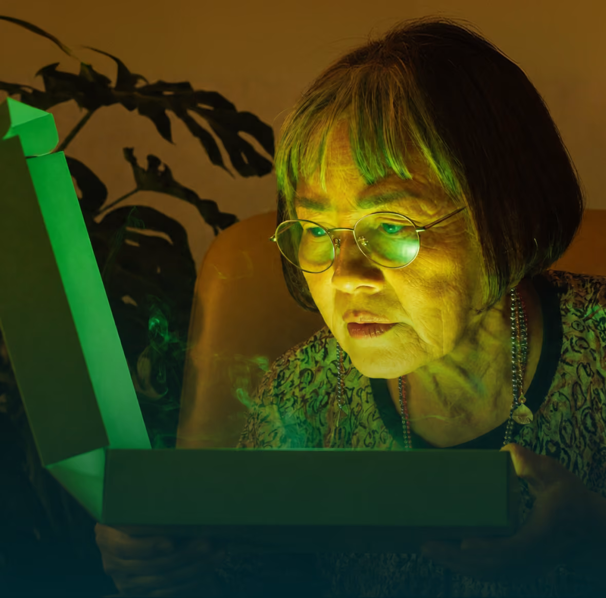 Older woman with glasses looking surprised and intrigued at an open glowing green box emitting light and smoke in a dimly lit room with a large plant in the background.