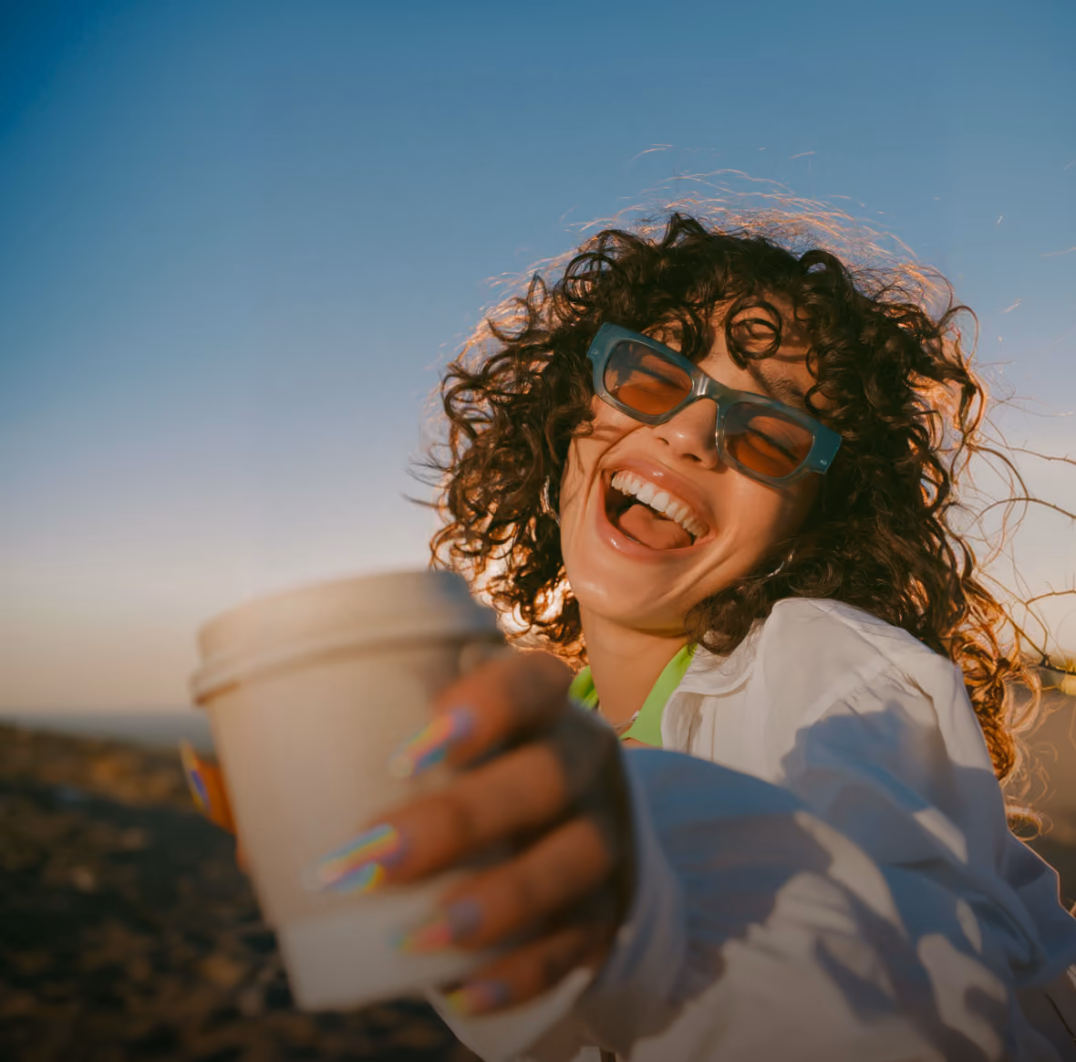Smiling young woman with curly hair wearing sunglasses and a white jacket, holding a takeaway coffee cup toward the camera outdoors at sunset.