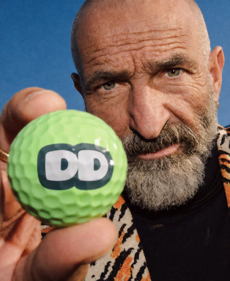 Close-up of a bearded man holding a bright green golf ball with the white and black 'DD' logo toward the camera, against a blue background.