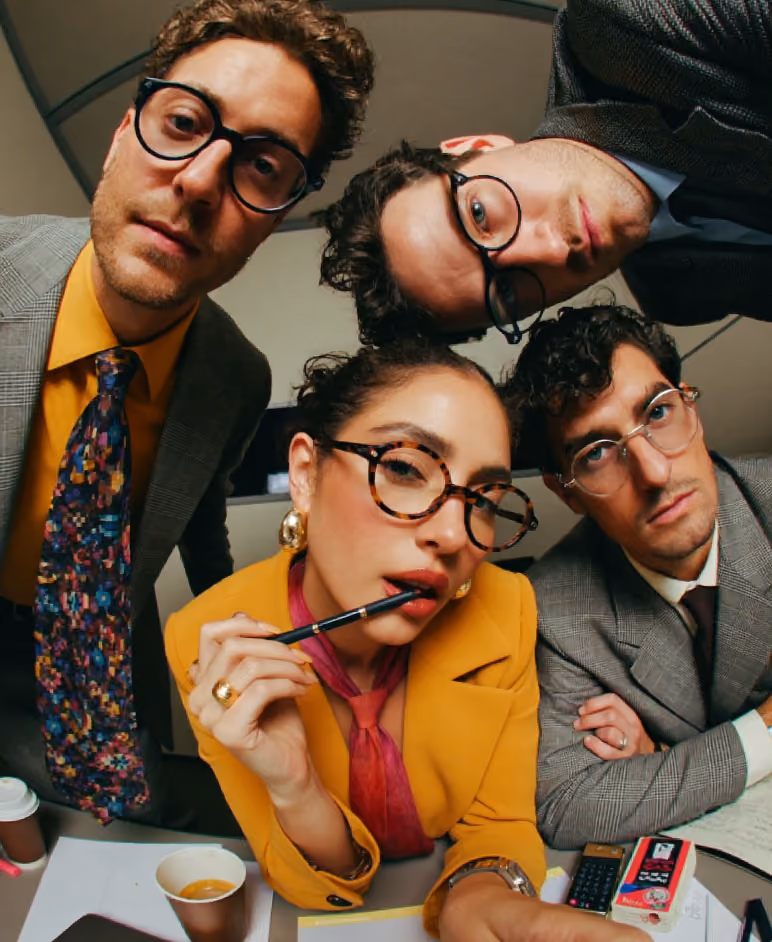 Four young adults wearing glasses, dressed in business attire, leaning toward the camera over a desk with coffee cups, documents, and a calculator, appearing engaged or contemplative.