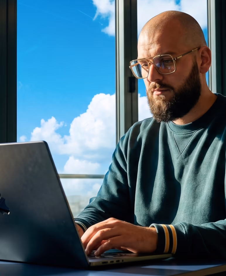 A bald man with a beard and glasses wearing a green sweatshirt is typing on a laptop by a window with a view of blue sky and clouds.