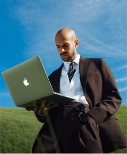 Man in a dark suit and tie standing outdoors on grass using an open Apple laptop under a clear blue sky.