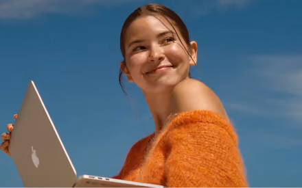 Smiling young woman wearing an orange fuzzy sweater holding an open laptop outdoors under a clear blue sky.