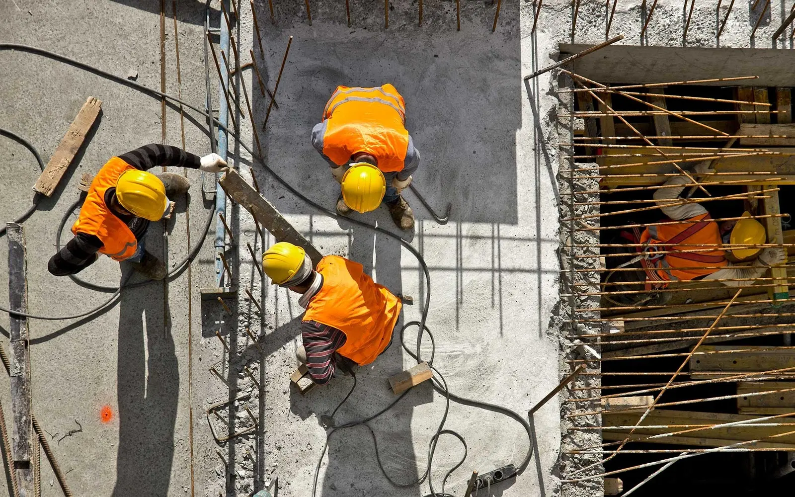 Construction workers in high-visibility jackets and hard hats tying steel reinforcement bars on a concrete floor slab at a heavy industry site, seen from above