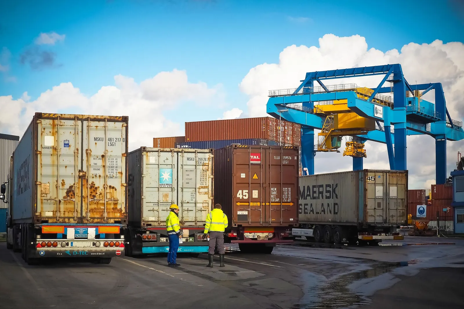 Two workers in hi-vis jackets checking paperwork beside container trailers at a logistics terminal, with a gantry crane and stacked shipping containers behind them