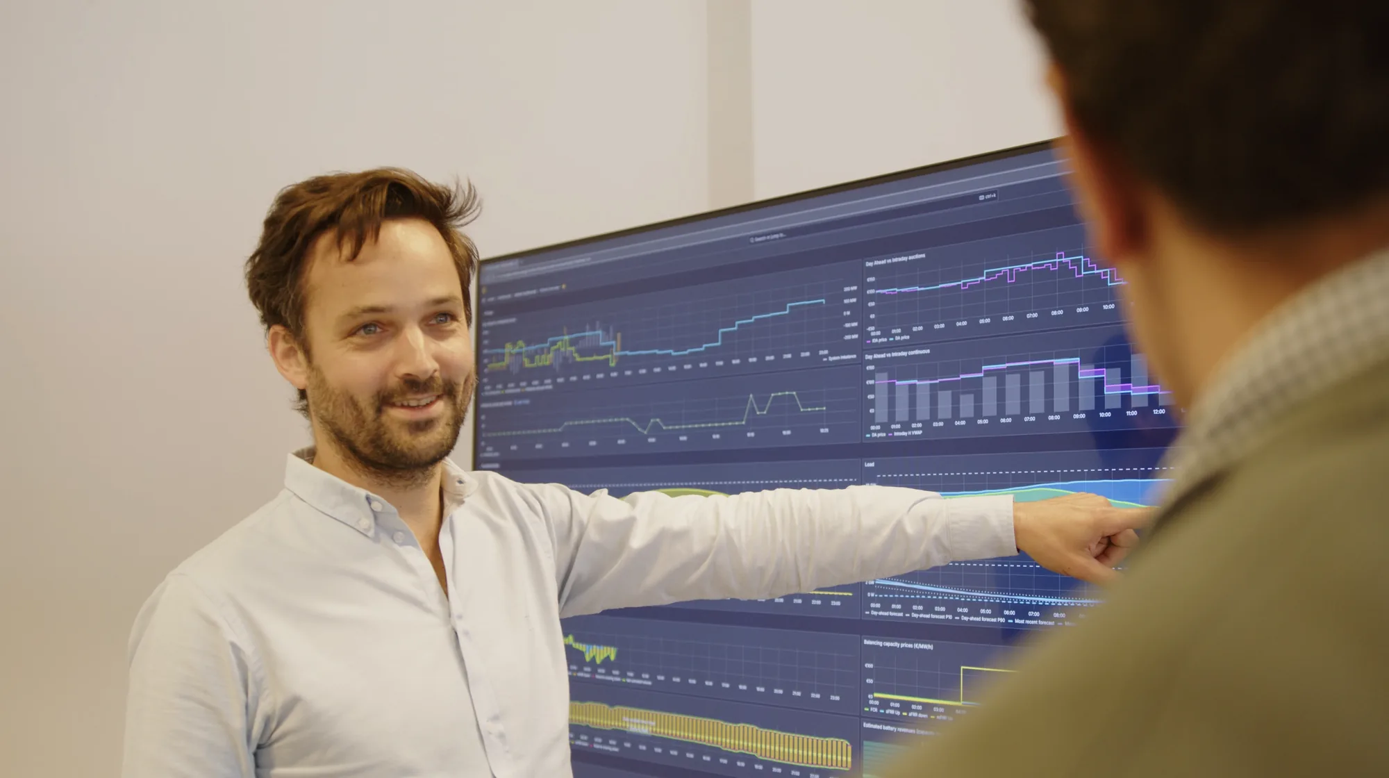 A Bnewable energy trader pointing at a live trading dashboard showing day-ahead and intraday market prices while explaining it to a colleague