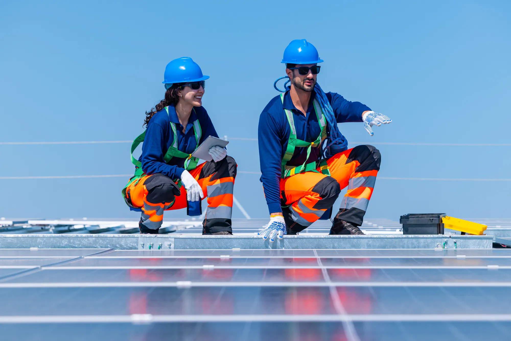 Two Bnewable technicians in hard hats and high-visibility clothing inspecting a rooftop solar installation with a tablet under a clear blue sky