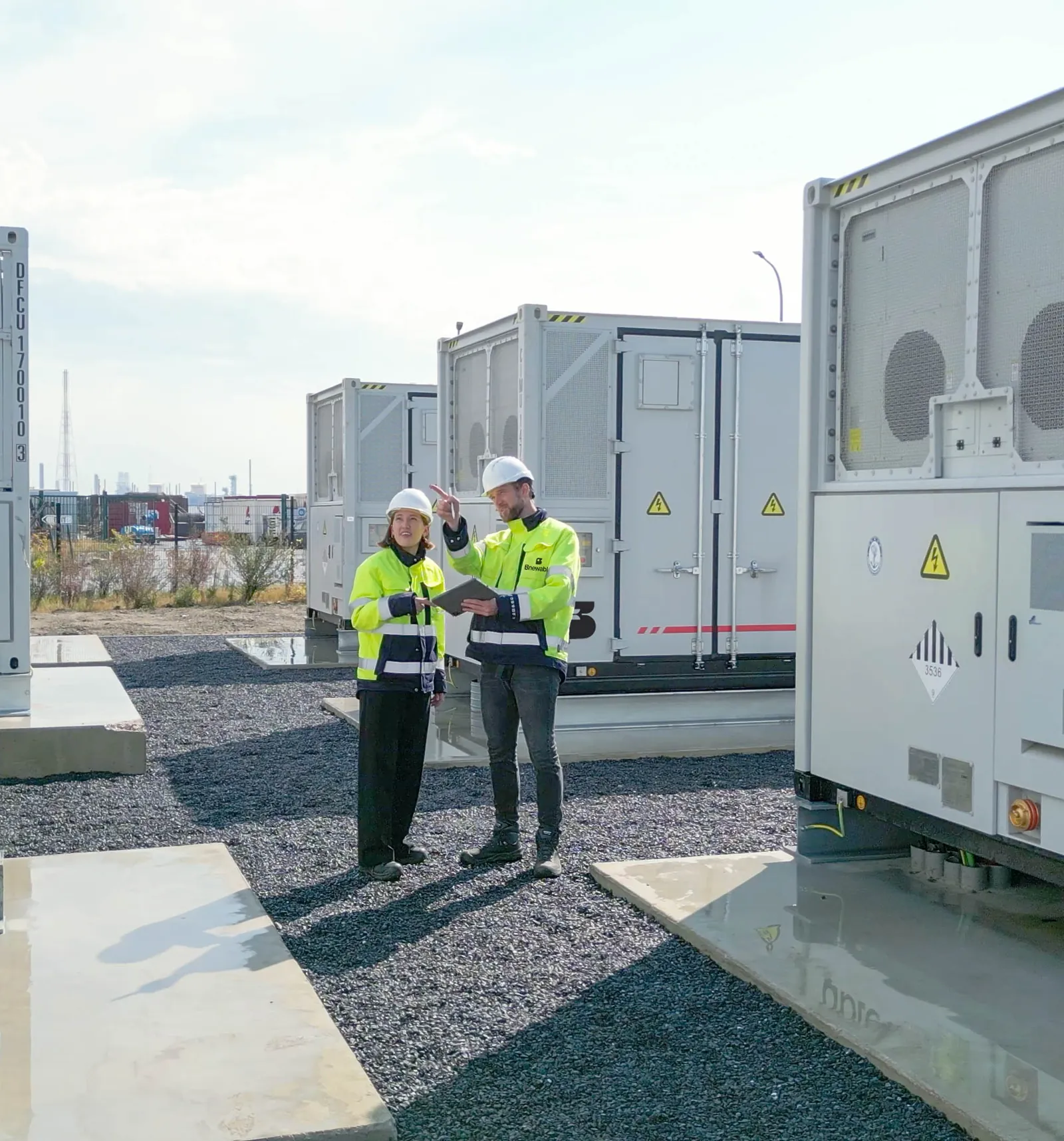 Two Bnewable engineers in high-visibility jackets reviewing data on a tablet between battery storage containers at an industrial site