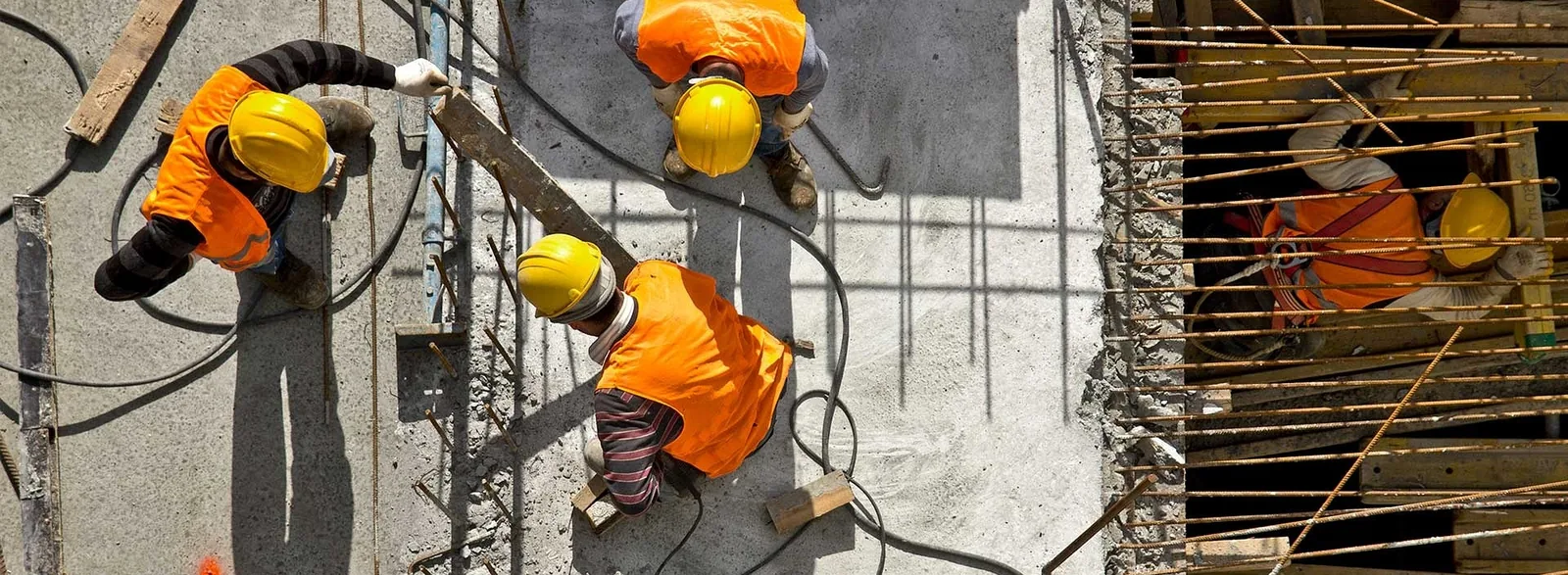 Construction workers in high-visibility jackets and hard hats tying steel reinforcement bars on a concrete floor slab at a heavy industry site, seen from above