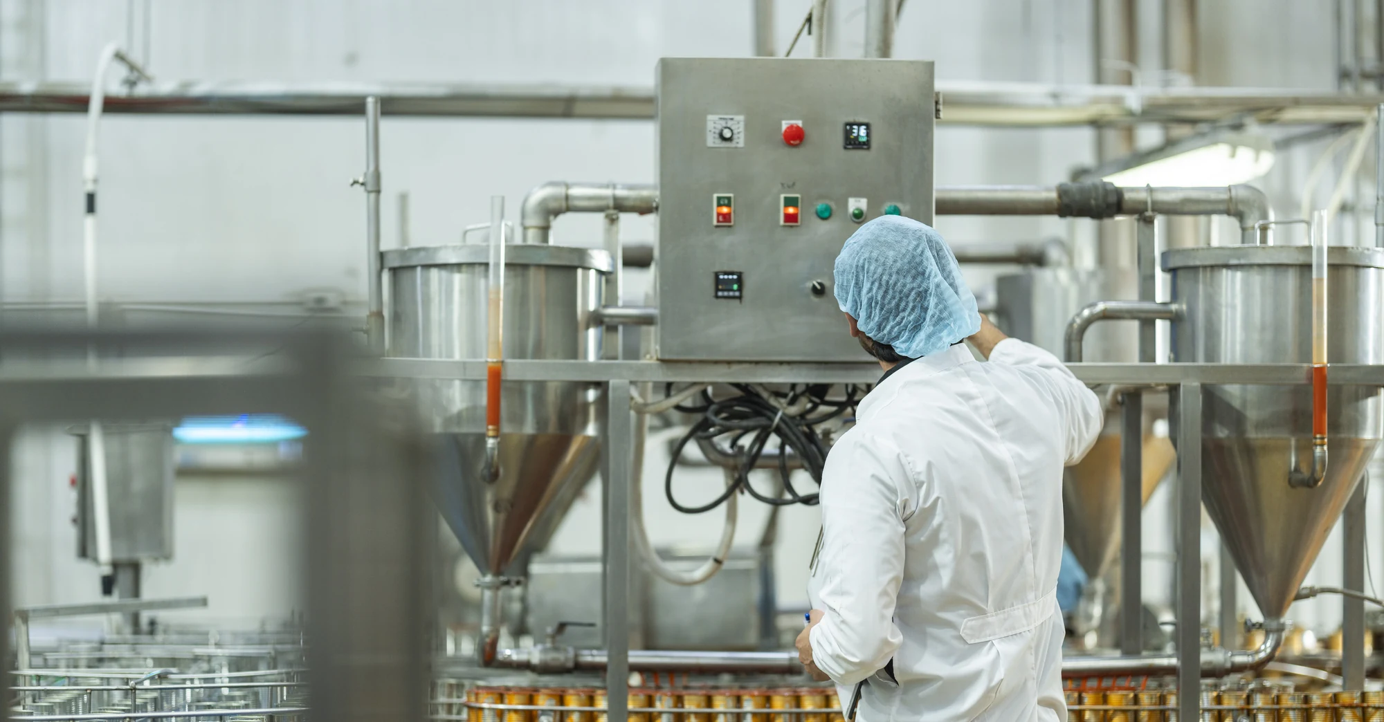 A worker in a white coat and blue hairnet checking a control panel in a food production facility, with stainless steel process tanks and a conveyor of filled cans