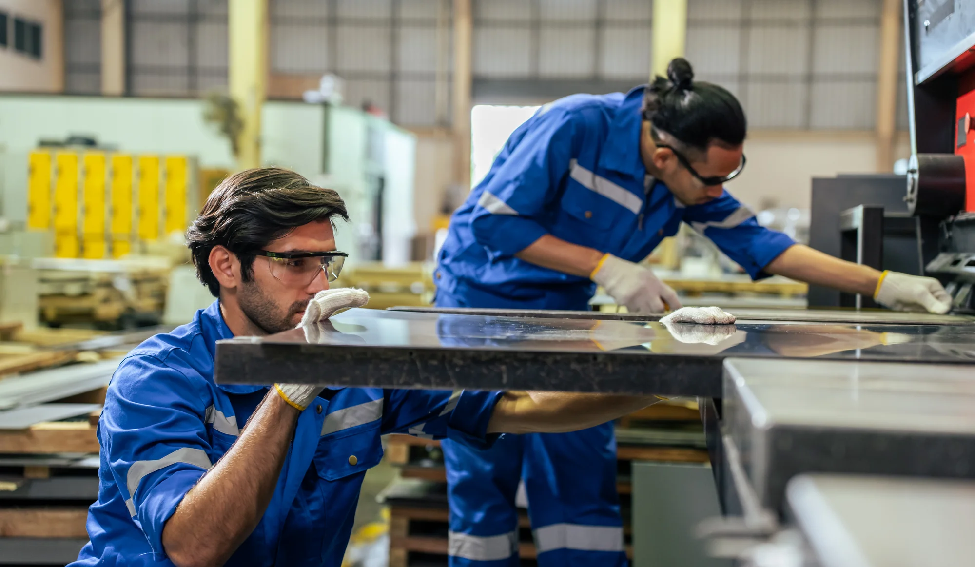 Two operators in blue workwear and safety glasses inspecting the surface of a polished metal sheet in a metalworking workshop, with a second operator working at a machine behind them