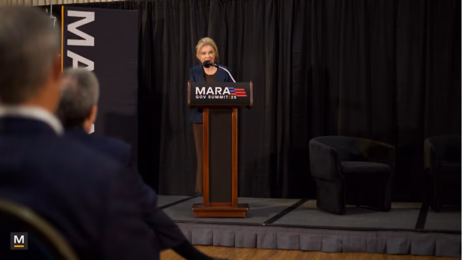 A woman speaking at a podium labeled 'MARA GOV SUMMIT 25' on a stage with black curtains and empty chairs, with an audience visible in the foreground.