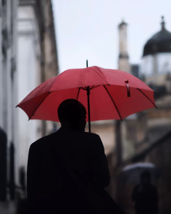 Silhouette einer Person mit dunkelrotem Regenschirm bei regnerischem Wetter in einer Stadt mit historischen Gebäuden im Hintergrund.