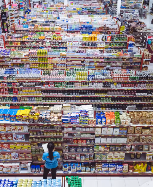 Blick aus der Vogelperspektive auf eine Person, die vor einem großen Supermarktregal steht, das prall gefüllt ist mit verschiedenen verpackten Lebensmitteln wie Frühstücksflocken, Snacks, Getränken und Süßigkeiten in bunten Verpackungen.