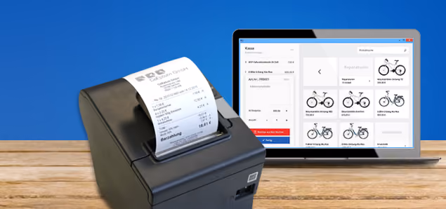 Schwarzer Belegdrucker mit einem Kassenbeleg vor einem Laptop auf einem Holztisch, auf dessen Bildschirm eine Fahrradverkaufs- und Reparatursoftware mit Produktbildern und Preisen zu sehen ist.