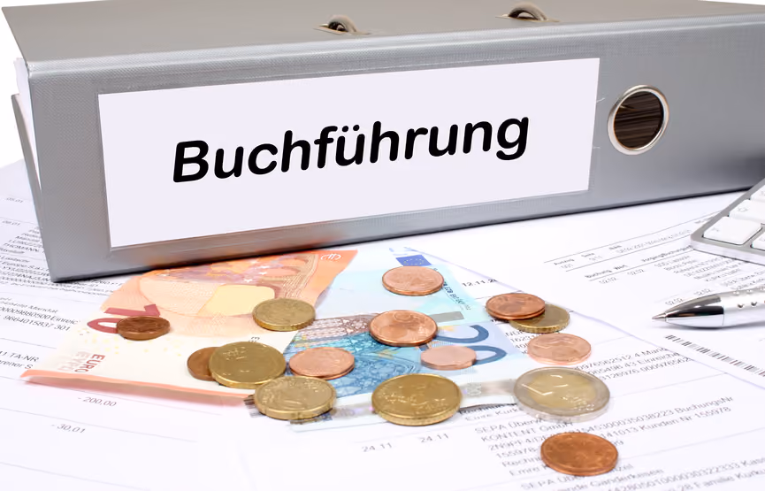 Ein grauer Ordner mit dem Etikett 'Buchführung' liegt auf Finanzdokumenten, daneben befinden sich Euro-Geldscheine, verschiedene Euro-Münzen, ein Stift und eine Computertastatur.
