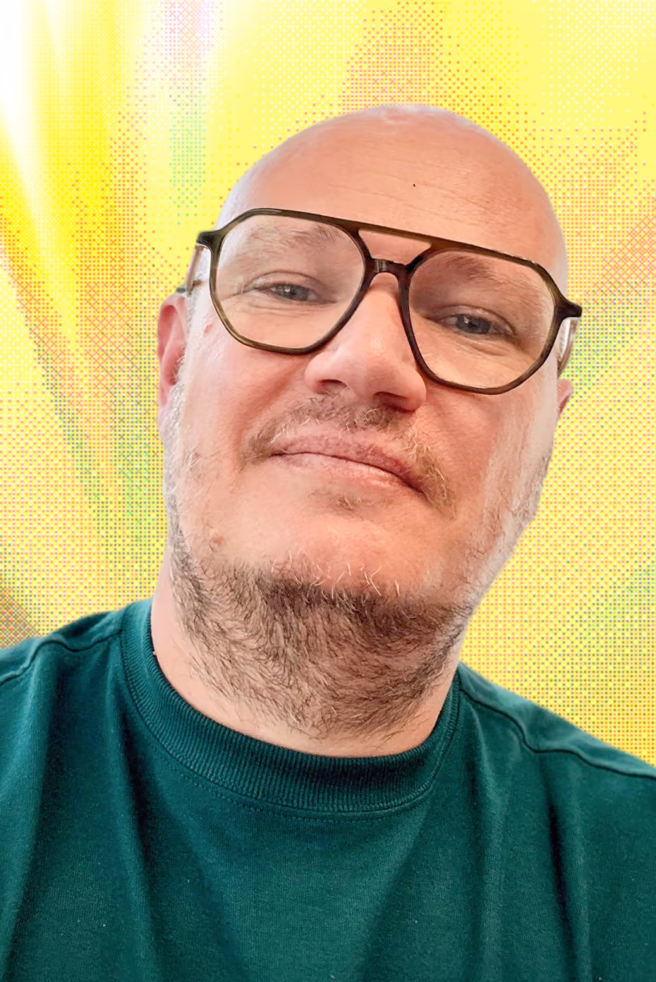 Homme chauve avec lunettes à monture noire portant un sweat vert devant un fond lumineux jaune.