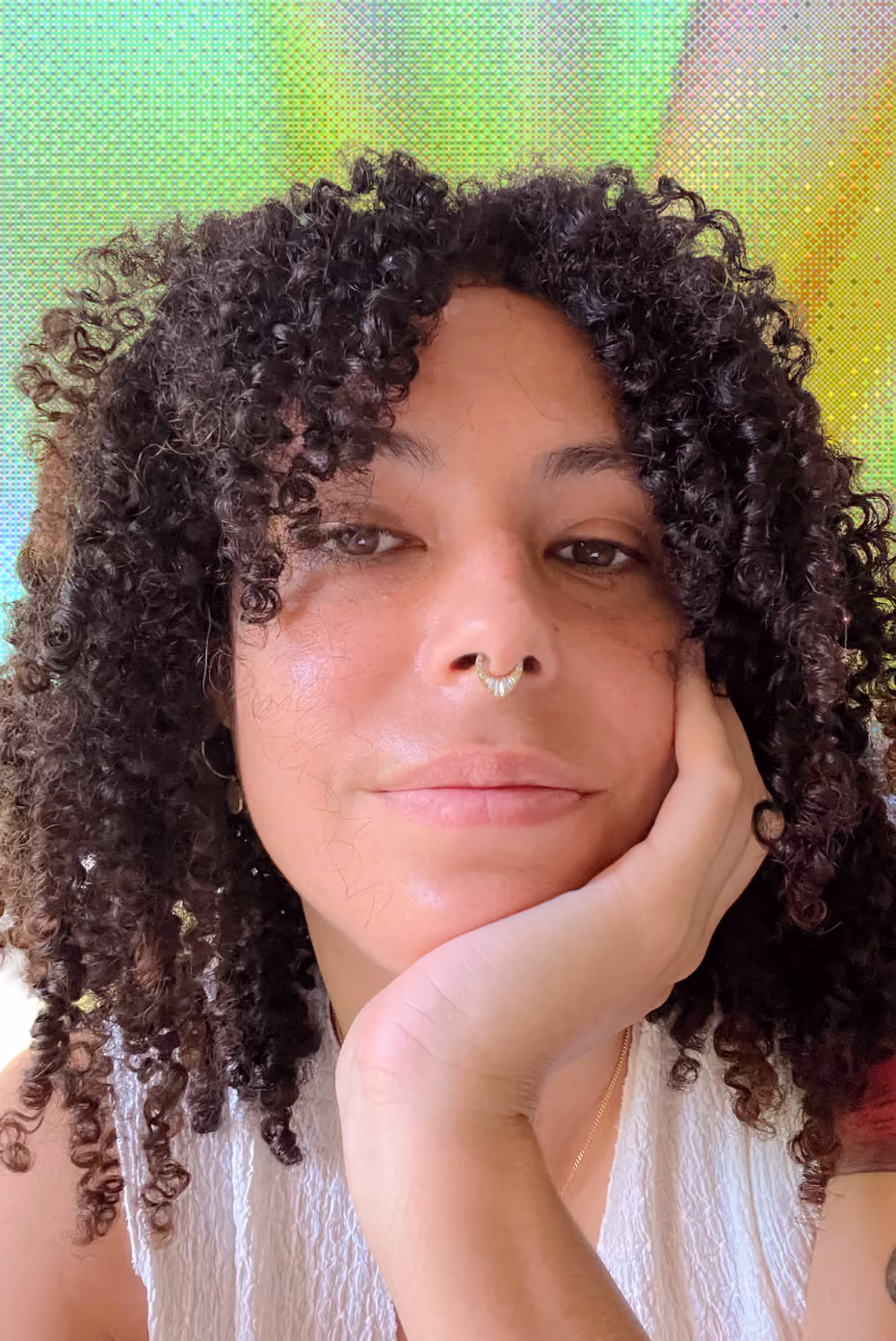 Portrait d'une personne avec des cheveux bouclés noirs, un septum doré et une expression détendue, posant la tête sur la main gauche, portant un haut blanc, avec un arrière-plan coloré aux tons verts et jaunes.