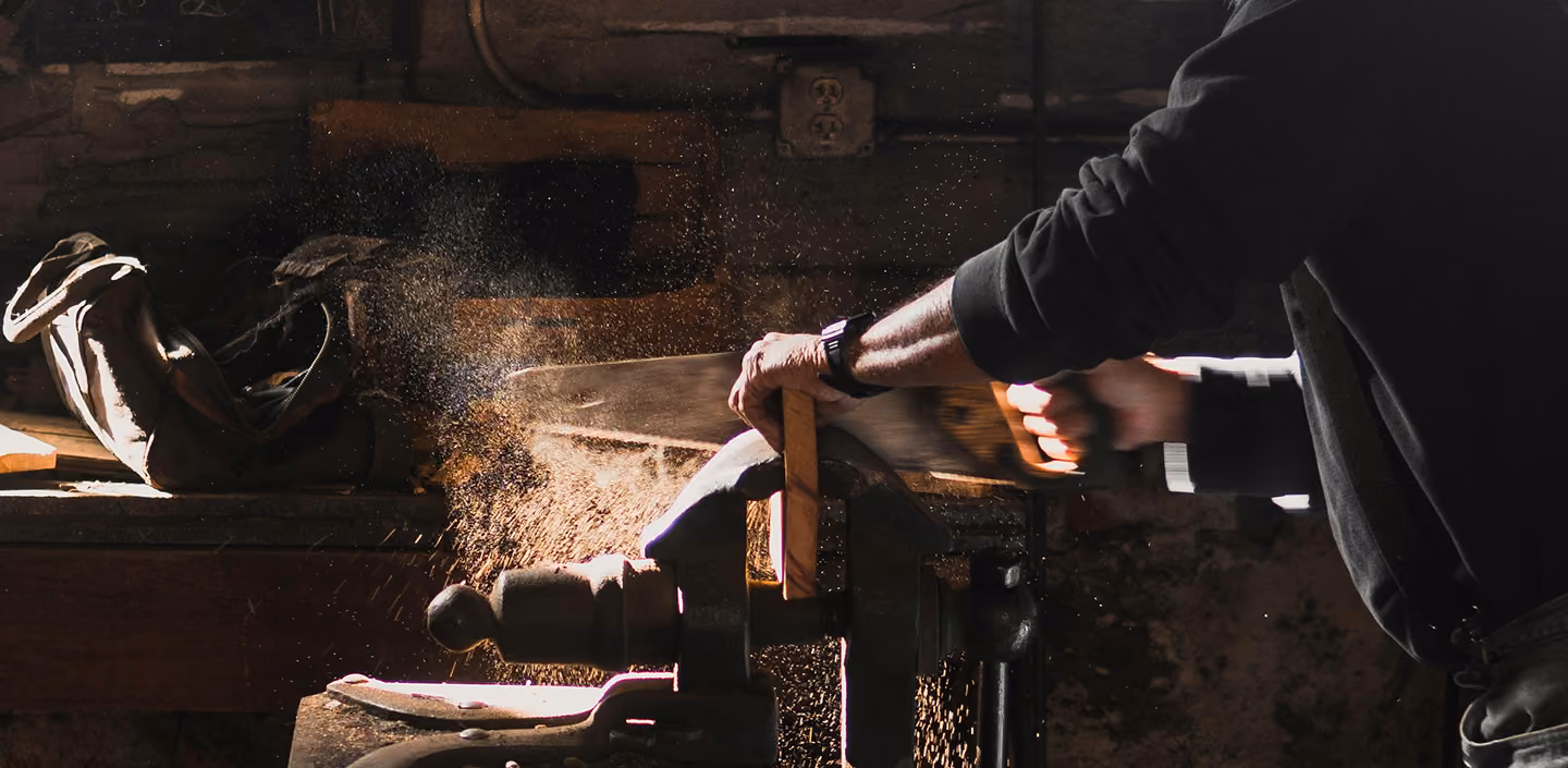 Person using a hand saw to cut wood clamped in a vise, with sawdust flying in a dimly lit workshop.