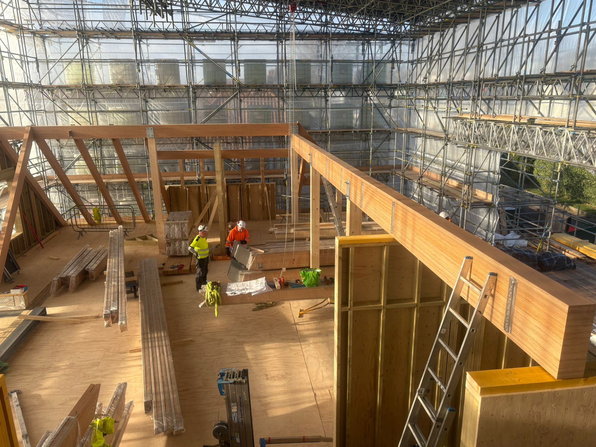 Indoor construction site with wooden framework and scaffolding surrounded by a translucent protective cover, showing two workers in safety gear handling materials on a wooden floor.