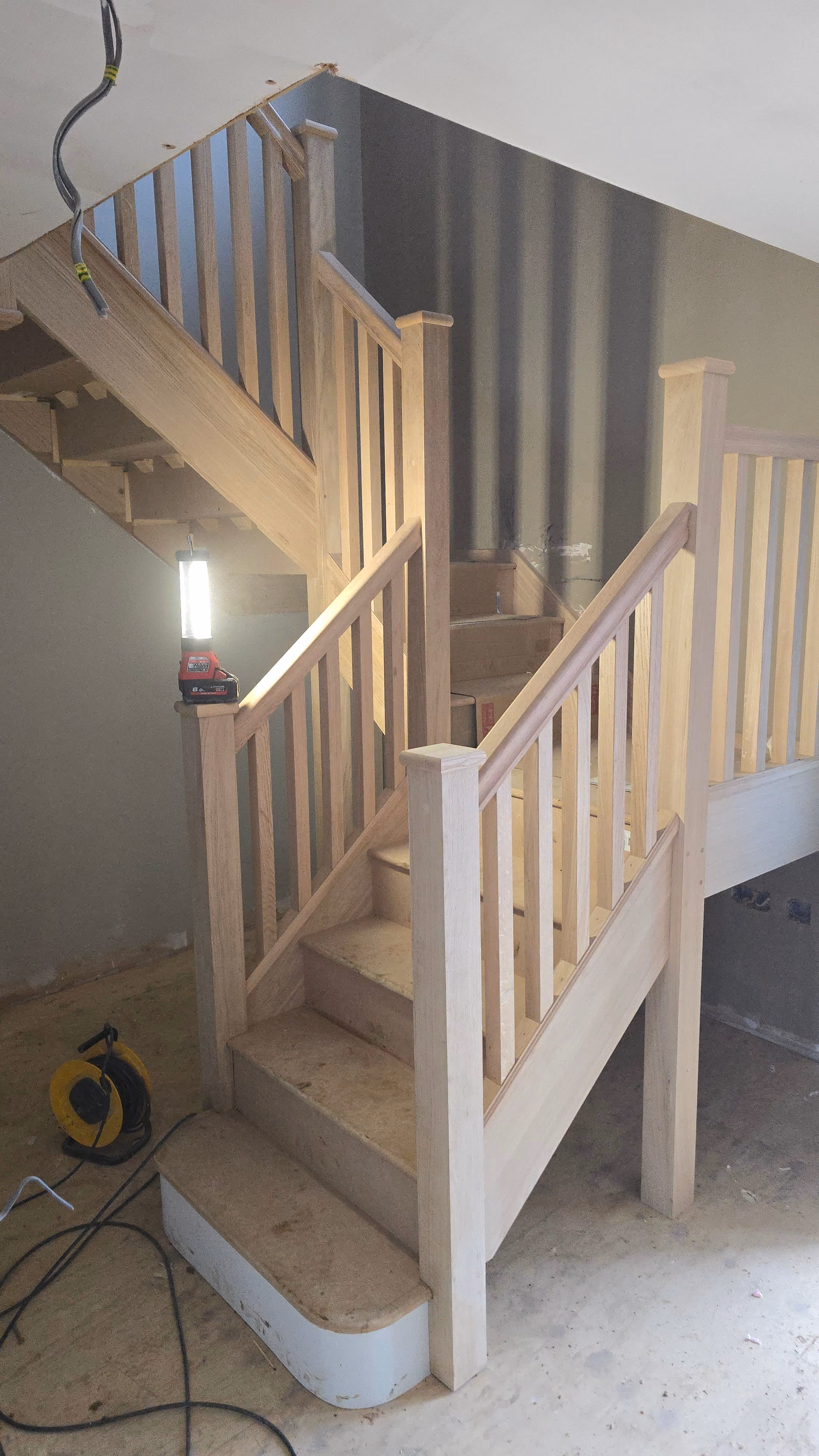 Partially constructed wooden staircase with railings and balusters, lit by a portable work light in a room under construction with exposed concrete floor and wall.