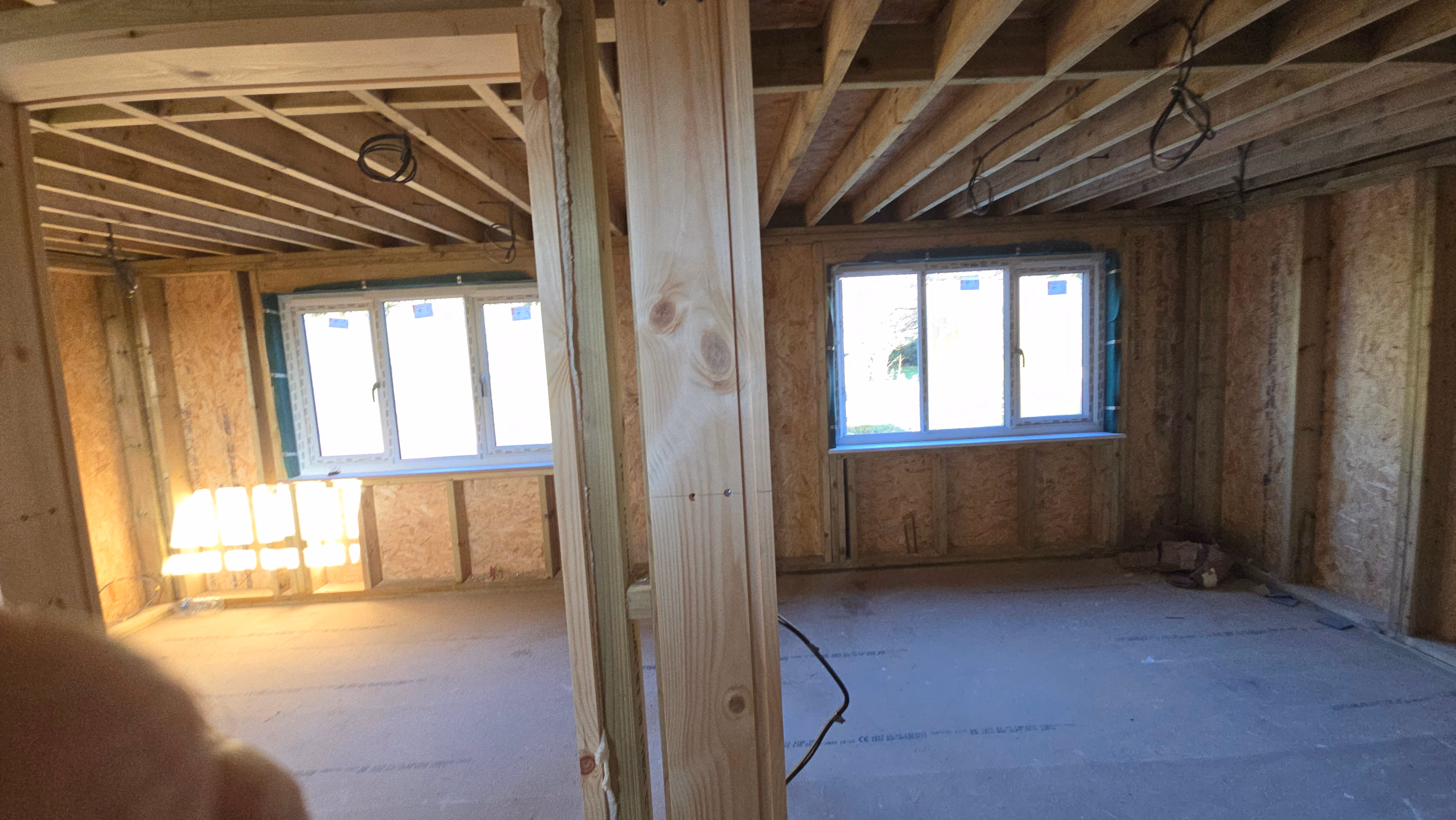 Interior of a wooden framed room under construction with exposed ceiling beams, two windows without trim, and exposed insulation walls.