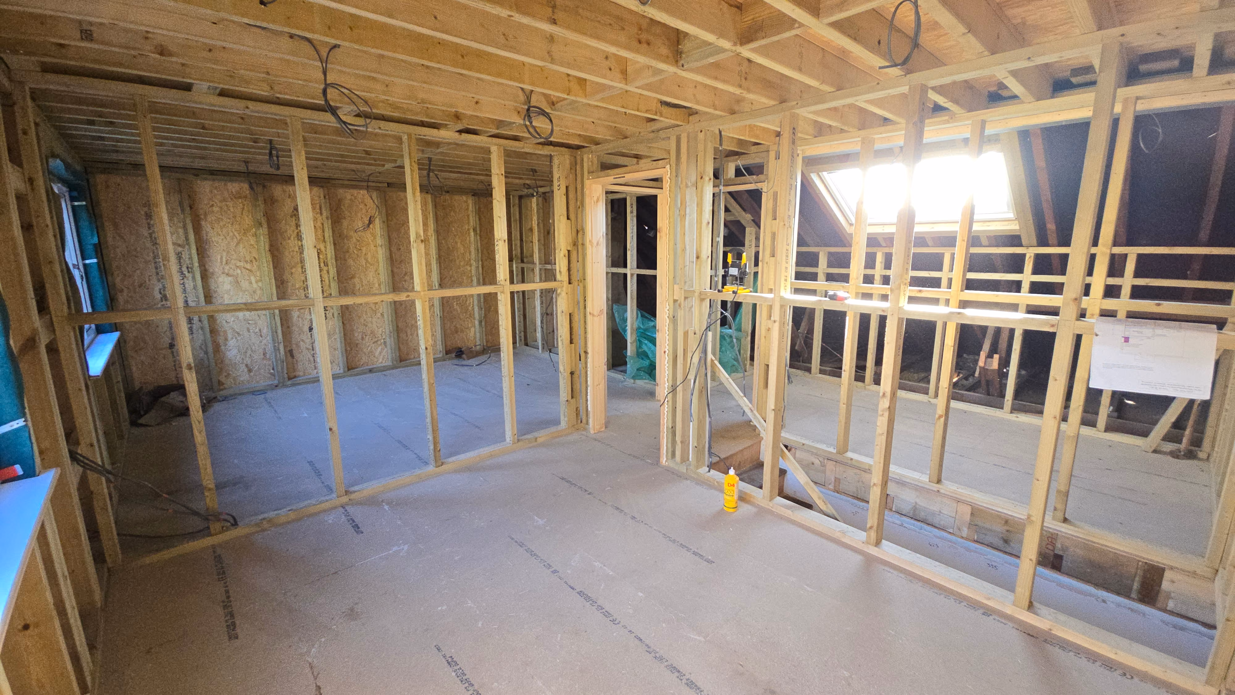 Interior view of a room under construction with exposed wooden framing, electrical wiring, and plywood floor and walls.