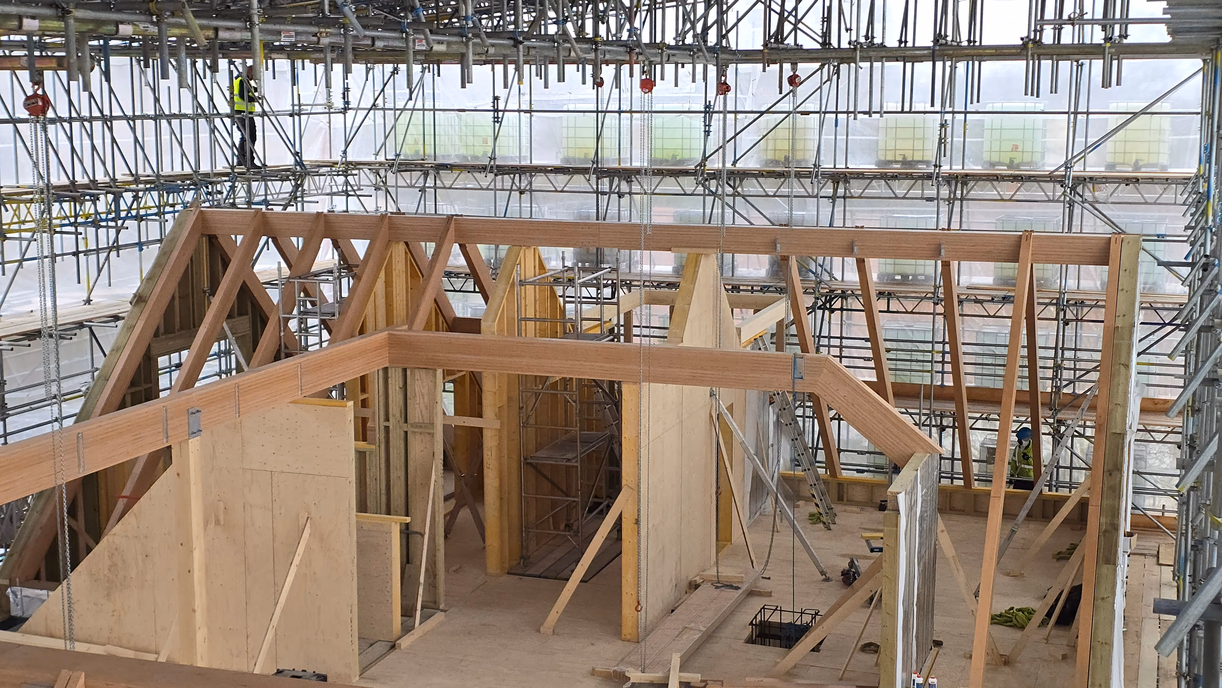 Interior view of a wooden house under construction with exposed wooden framing and prefabricated panels surrounded by metal scaffolding and a high ceiling structure.