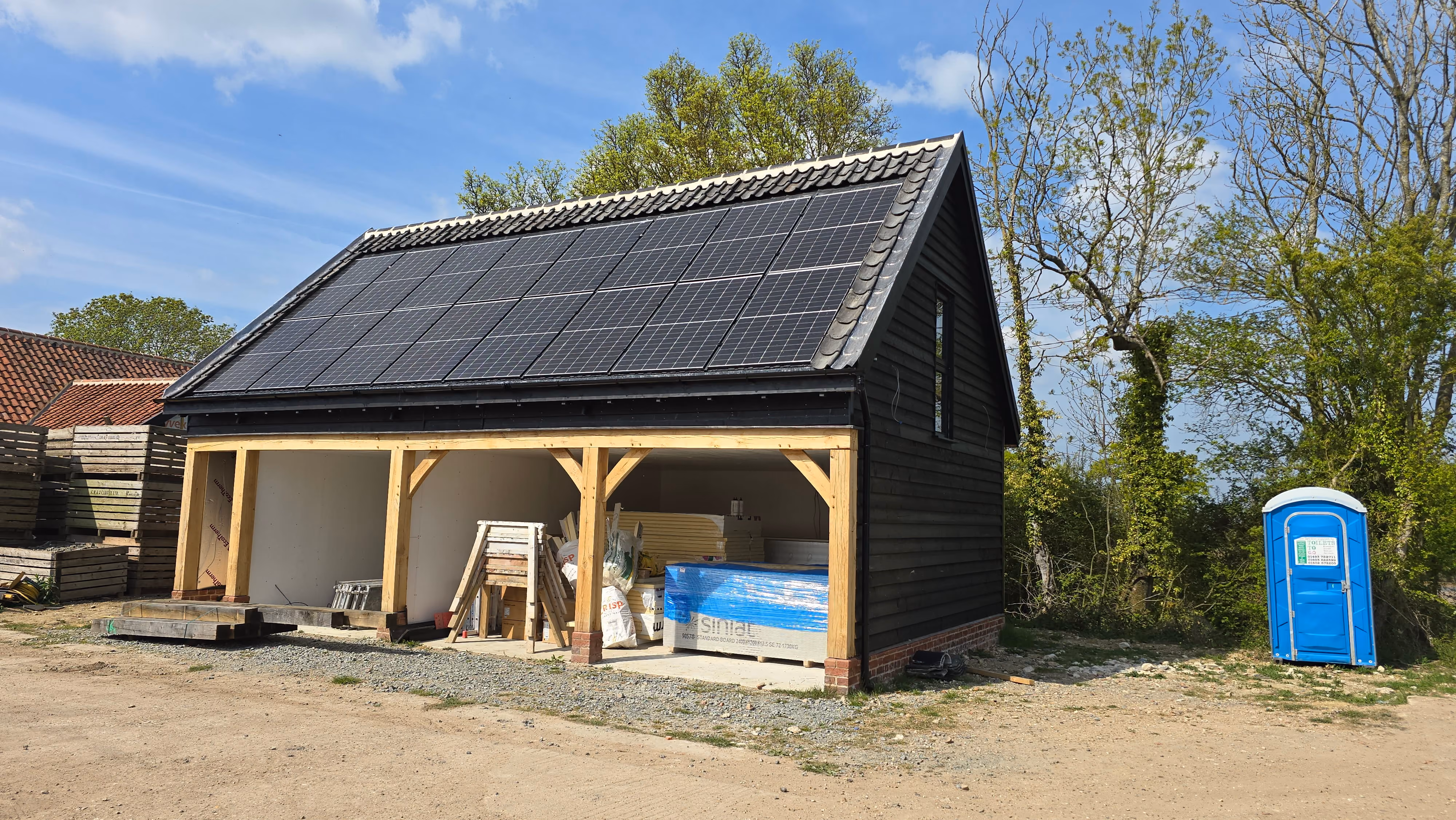 A small black wooden building with a slanted roof covered in solar panels, an open front section storing construction materials, surrounded by dirt ground and trees, with a blue portable toilet nearby.
