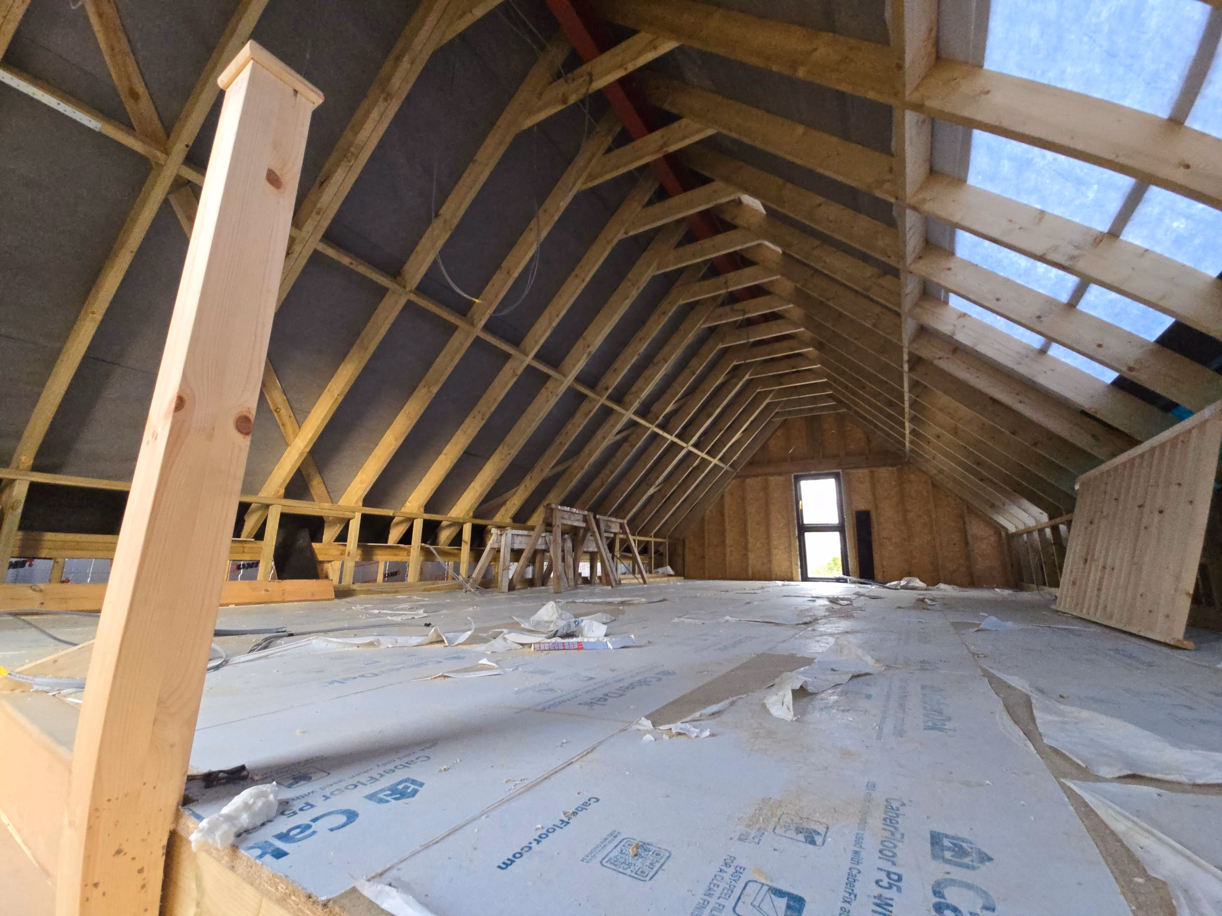 Interior of an attic under construction featuring wooden framing, large floor insulation panels, and a window at the far end letting in light.