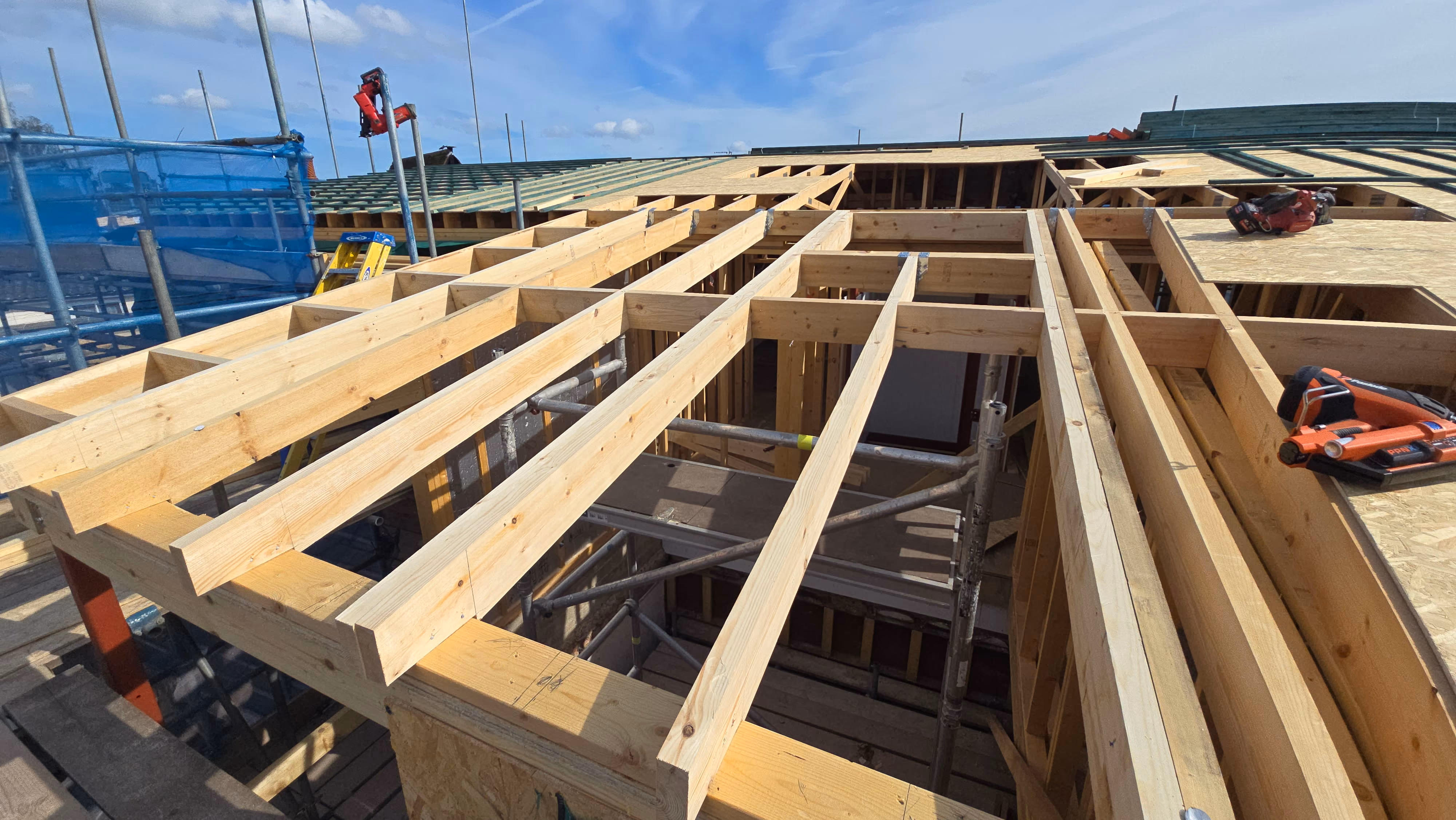 Wooden roof framing with exposed beams on a building under construction, scaffolding inside, and tools on plywood sheets partially covering the structure under a blue sky.