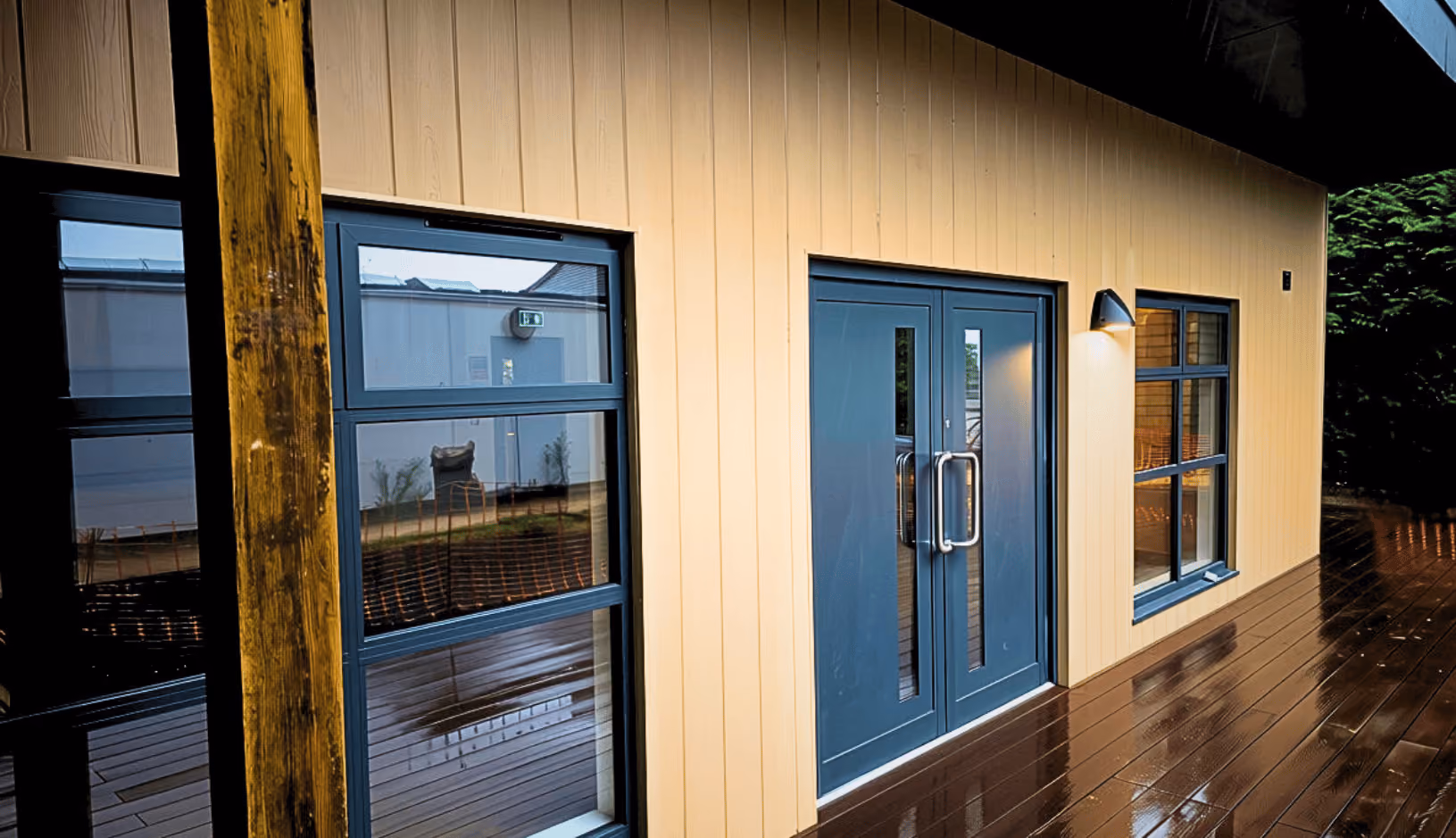 Exterior view of a building with beige vertical siding, a dark blue double door with glass panels, adjacent windows with blue frames, a wooden post, and a glossy wooden deck reflecting light.
