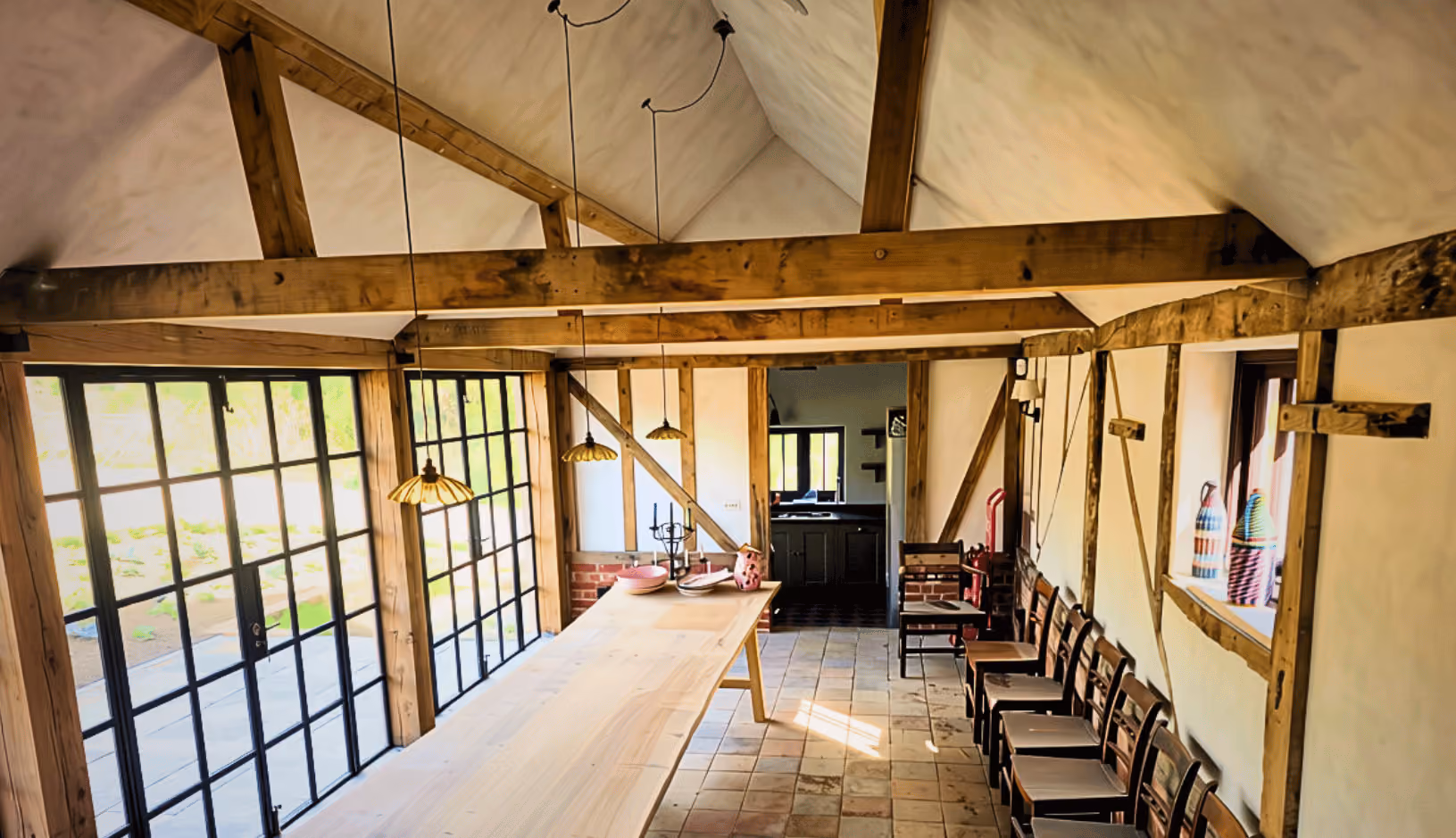 Interior of a rustic dining area with wooden beams and trusses, a long wooden table, hanging pendant lights, tiled floor, and multiple chairs lined up against the right wall under a large window.