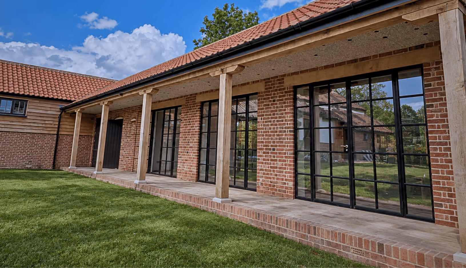 Long brick building with a tiled roof supported by wooden pillars, featuring large glass doors and windows overlooking a green lawn under a partly cloudy blue sky.