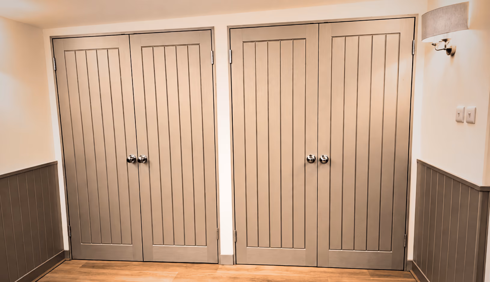 Interior hallway with two sets of beige double doors featuring vertical panels and silver round handles, wooden floor, white walls with gray wainscoting, and a wall-mounted light fixture on the right.