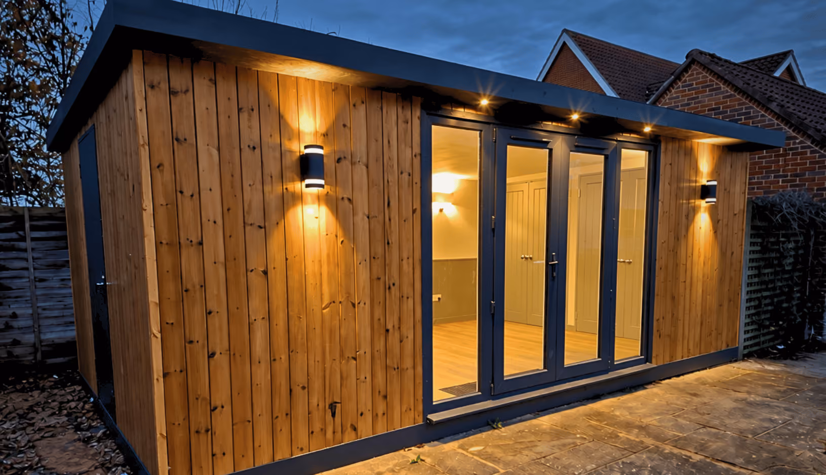 A modern wooden garden room with vertical timber cladding and black trim, illuminated by exterior wall lights at dusk, with large glass double doors showing a lit interior.