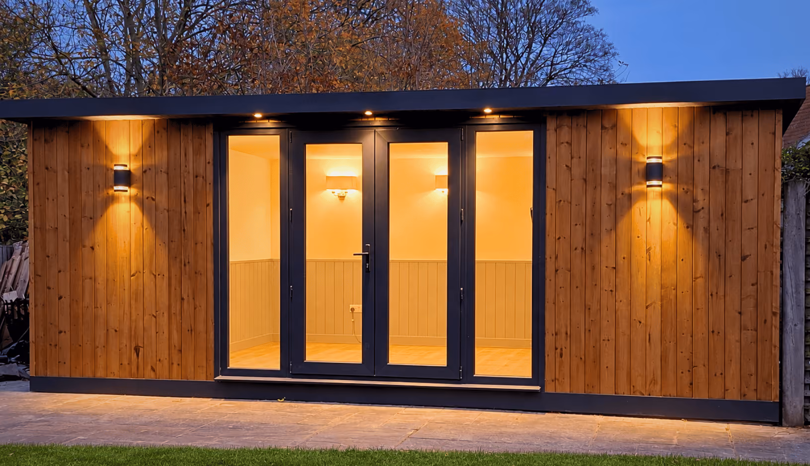 Modern wooden garden office with black-framed glass double doors, warm interior lighting, outdoor wall lights, and a paved path in front with grass.