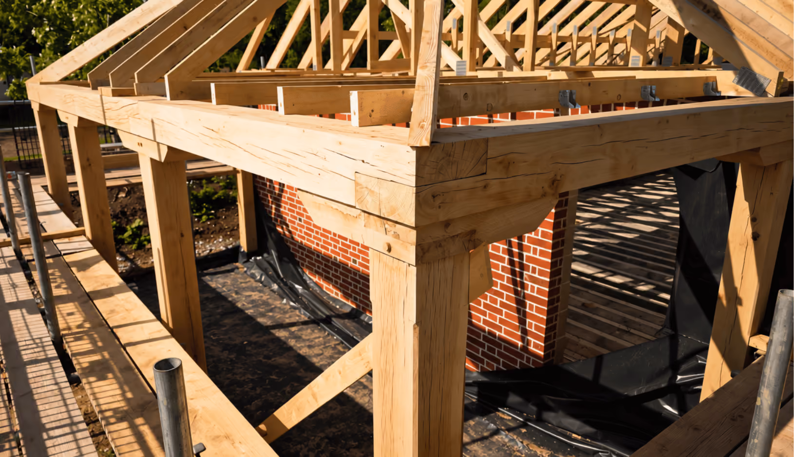 Wooden framework of a building under construction with visible beams and a brick wall in the background.