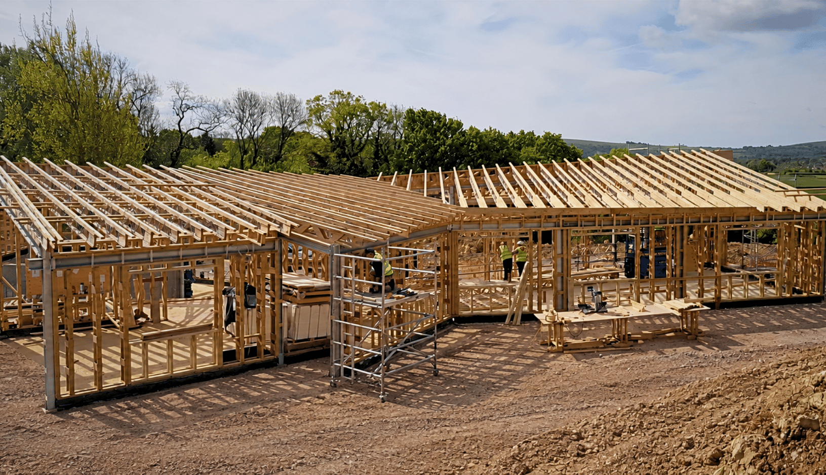 Wooden framework of a large building under construction on a dirt site with scaffolding and workers wearing yellow safety vests, surrounded by green trees and a partly cloudy sky.