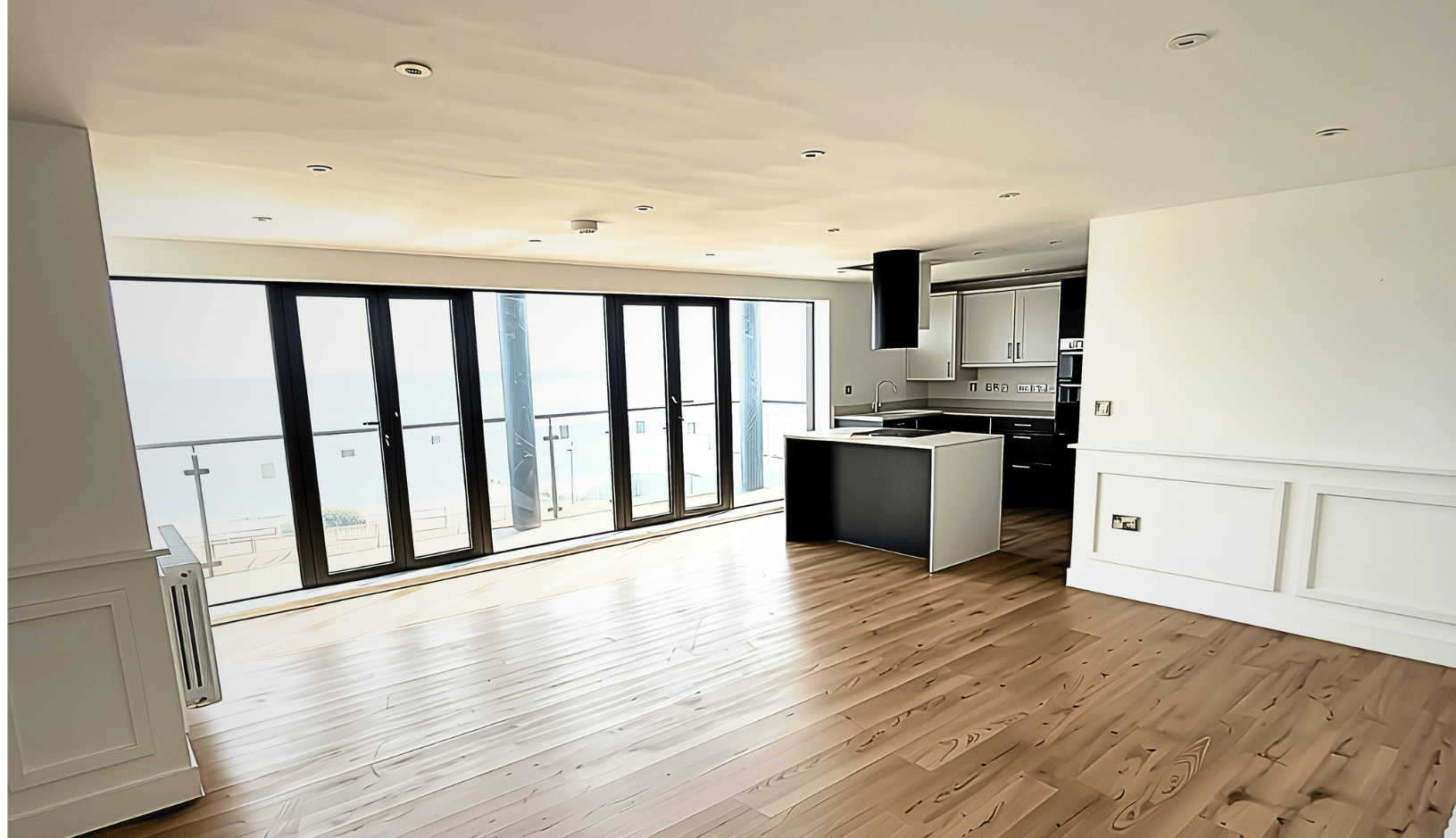 Bright modern open-plan kitchen and living area with wooden flooring, white cabinetry, a central island, and large floor-to-ceiling glass doors overlooking a balcony with a sea view.