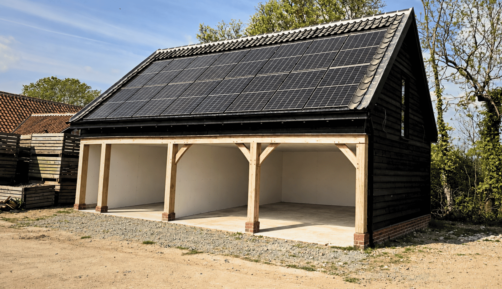 Garage with three open bays, a wooden frame, dark siding, and a roof covered in solar panels under a clear sky surrounded by trees and gravel ground.