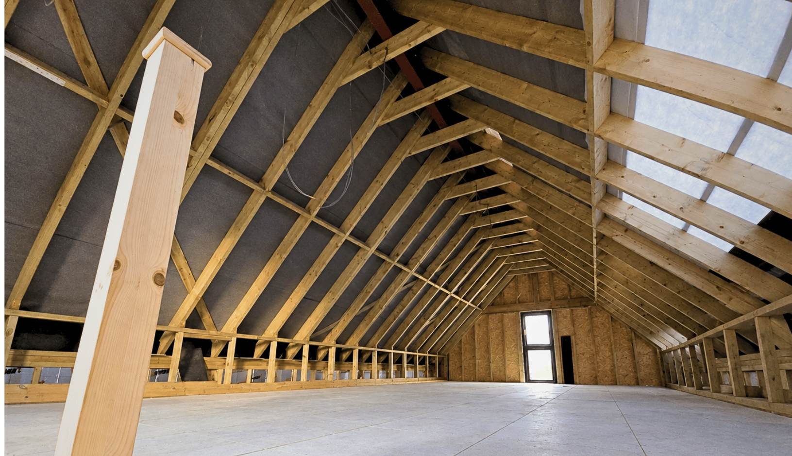 Spacious unfinished attic with wooden beams forming a pitched roof structure, plywood flooring, and a door-sized window at the far end letting in natural light.
