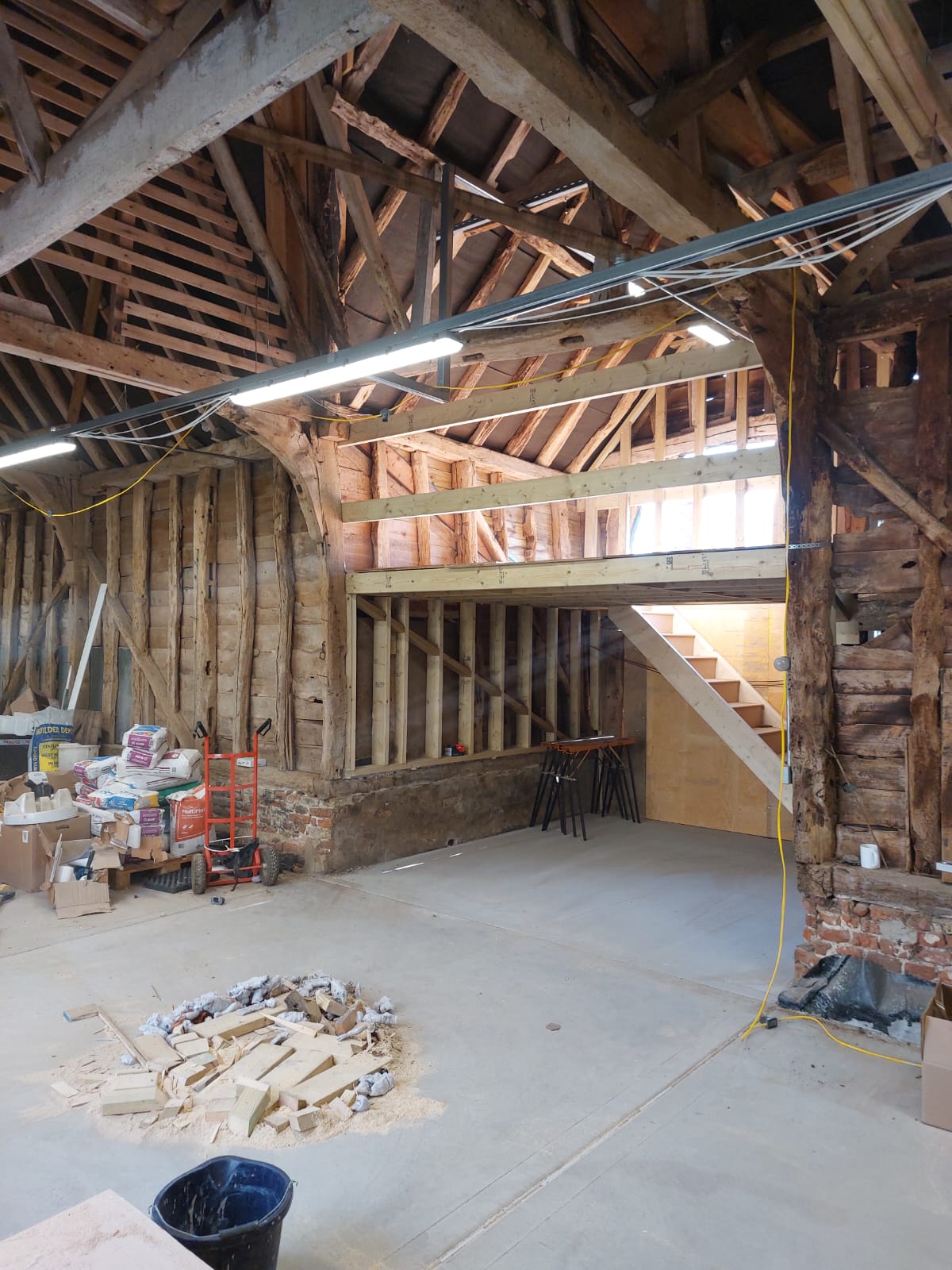 Interior of a wooden barn or workshop under renovation with exposed wooden beams and framework, a pile of wood scraps on a concrete floor, construction materials stacked in one corner, and a staircase leading to a loft area.