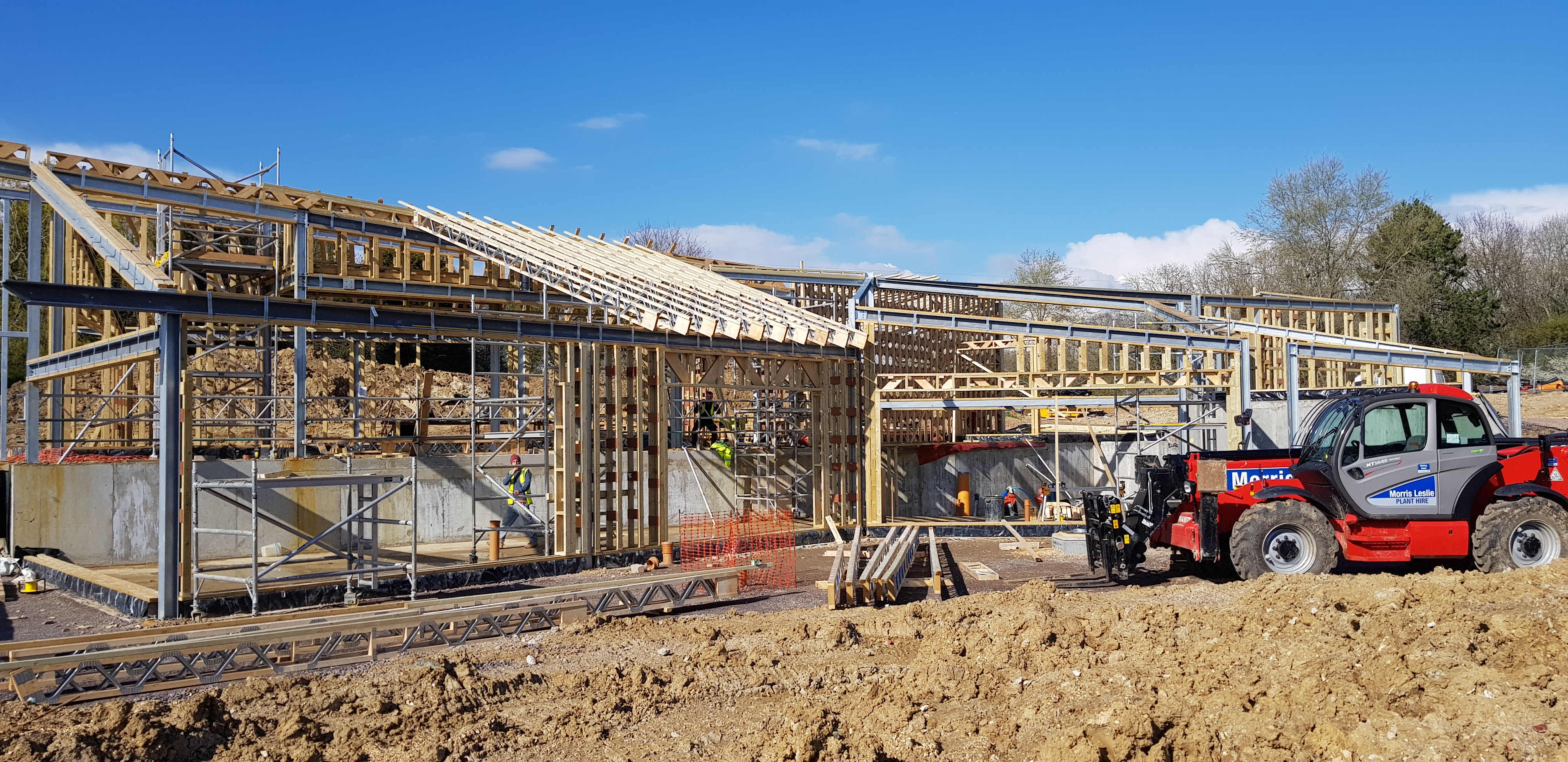 Construction site with wooden framing and metal supports for a building under clear blue sky, workers in safety gear present, and a red telehandler vehicle labeled Morris Leslie on the right side on dirt ground.