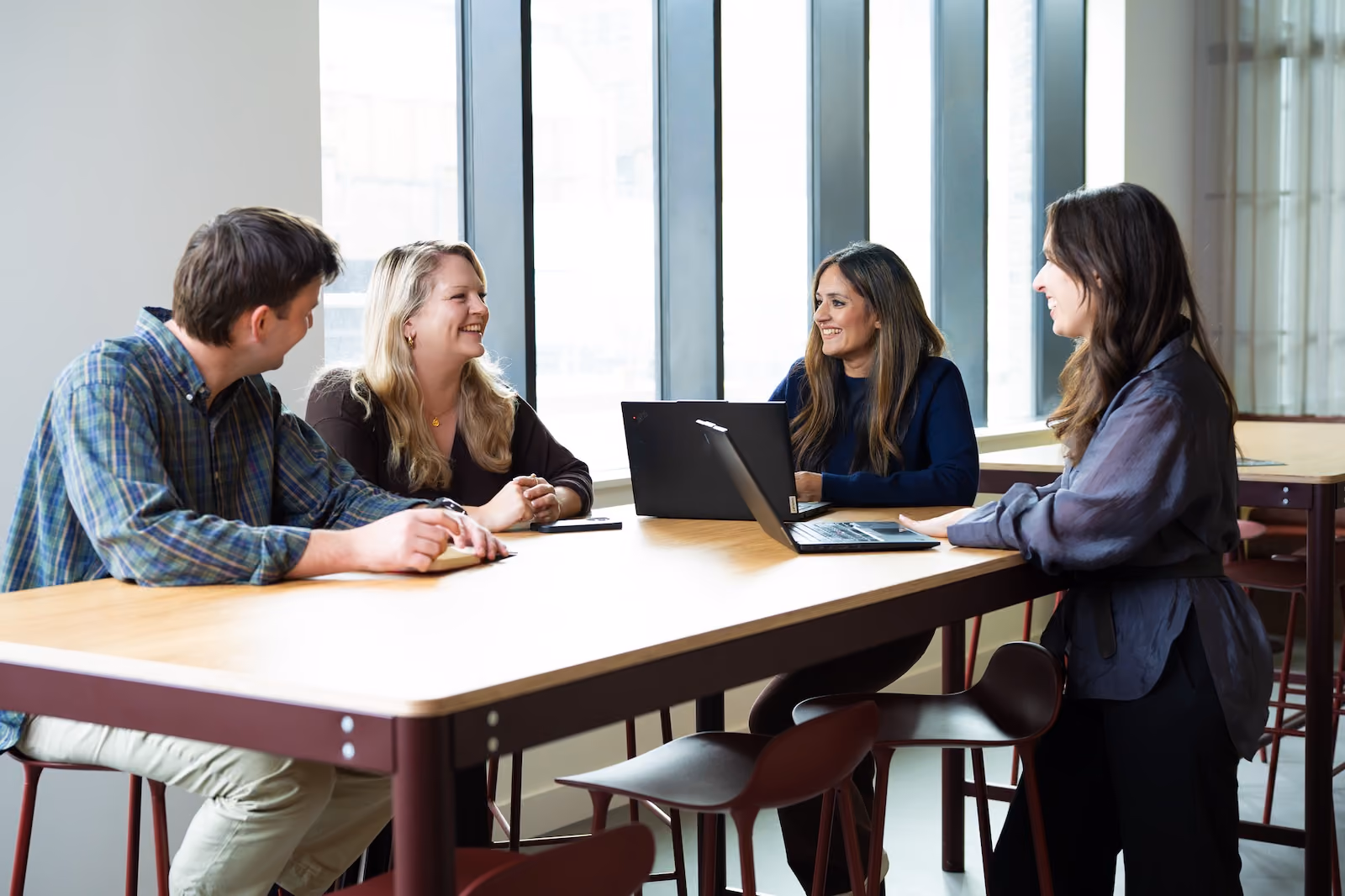 Four people sitting and standing around a wooden table, engaged in conversation with two open laptops in a bright room with large vertical windows.