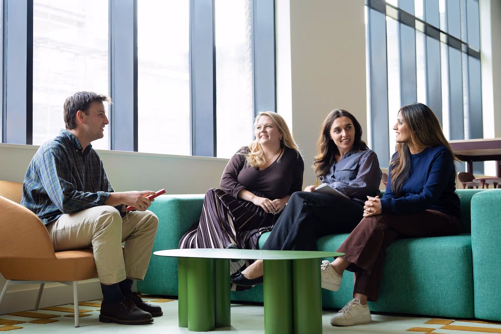 Four young adults having a friendly conversation seated in a modern lounge with large windows; one man sits in an armchair holding a red phone, three women sit on a green couch engaged and smiling.