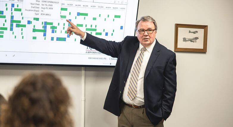 Tom Quinn pointing at a screen displaying a colorful bar chart during a presentation, with a blurred person in the foreground and a framed picture of airplanes on the wall.