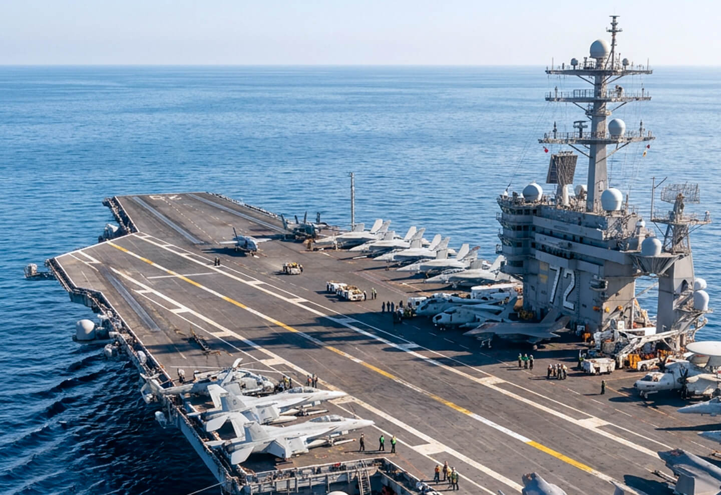 Aircraft carrier on calm sea with multiple fighter jets and helicopters parked on the flight deck, and crew members standing beside aircraft and along the deck. The ship's island structure displays the number 72.